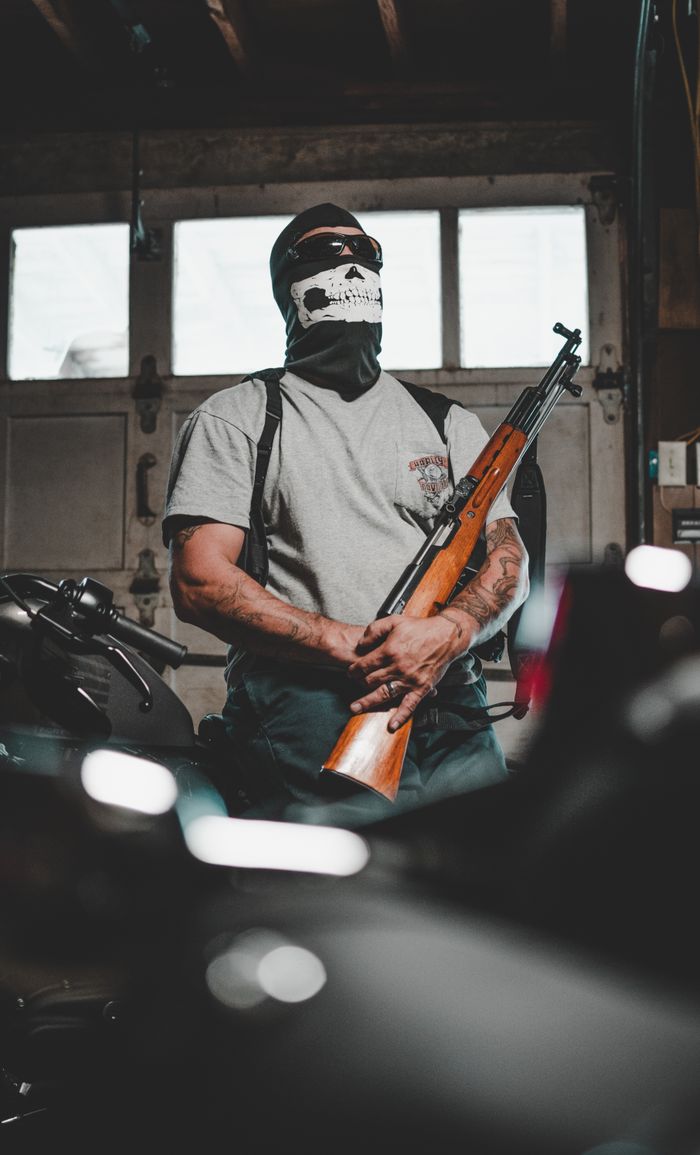 about-01 A masked person stands in a garage holding a rifle, surrounded by motorcycles.