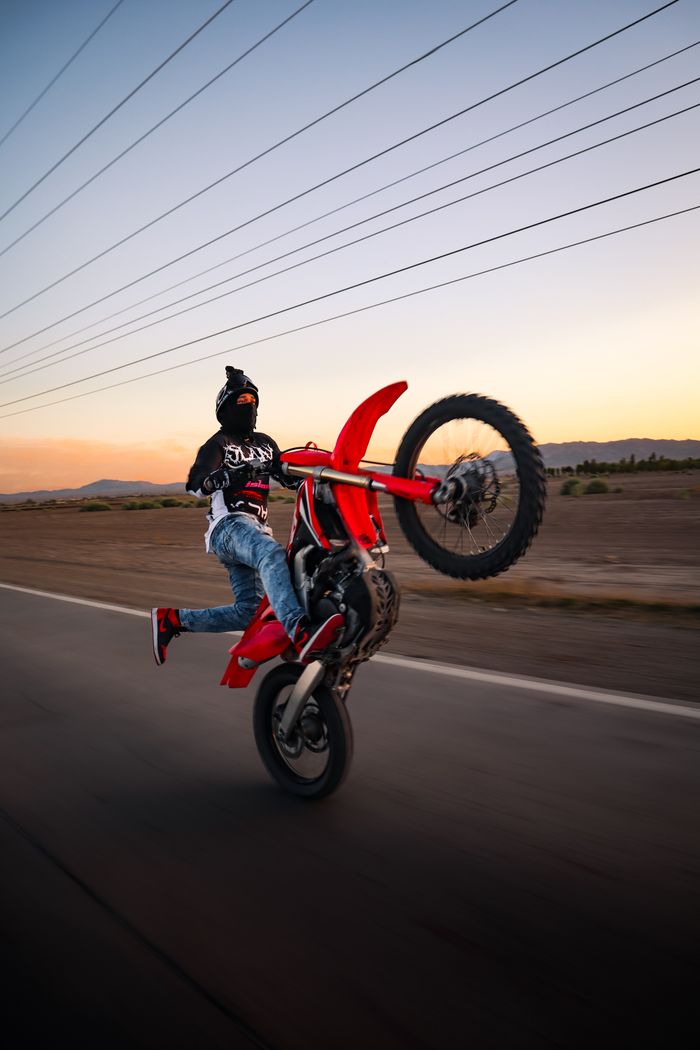 gallery-04 Thrilling stunt of a motorcyclist executing a wheelie on a deserted road during a stunning sunset.