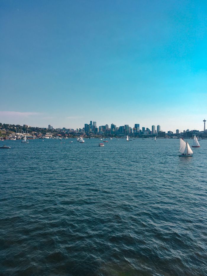 about-02 Serene view of sailboats on a blue lake with a city skyline in the distance.